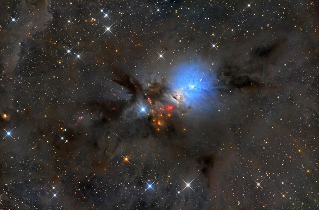 Mitten im Bild leuchtet ein blauer Reflexionsnebel. Er ist von dunklen Staubwolken umgeben. Rechts unter dem blauen Nebel sind kleine rote Lichter, es sind Anzeichen von Sternbildung.