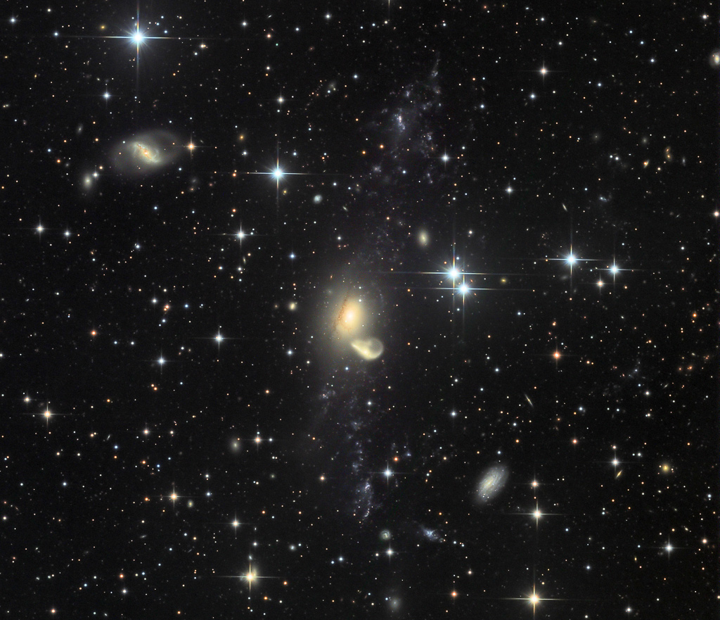 Mitten im Bild kommen zwei Galaxien einander sehr nahe. Die kleinere Galaxie links unten ist muschelförmig. Nach oben und unten zieht sich ein Gezeitenschweif hinaus, in dem viele Zwerggalaxien entstehen. Das Bildfeld ist voller Sterne und Galaxien.