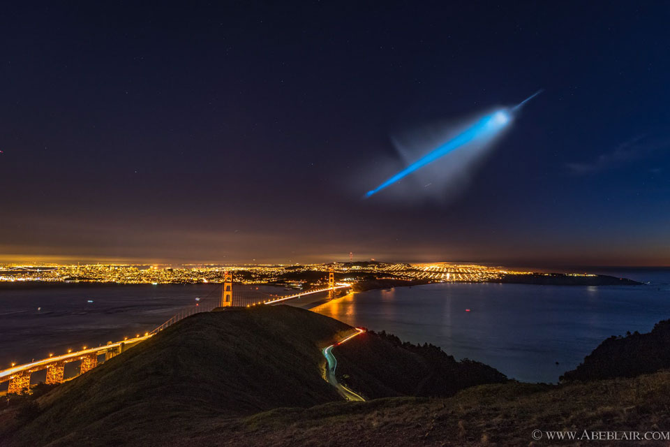 Über San Francisco breitet sich am dunklen Himmel eine blau leuchtende Abgasfahne aus. Vorne liegen die Bucht und die Golden Gate Bridge.