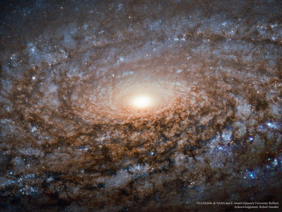 Um ein helles Galaxienzentrum wirbeln wild strukturierte Spiralarme voller Dunkelnebel und fleckiger blauer Sternhaufen.