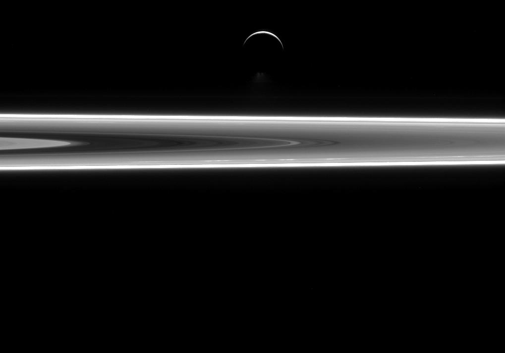 Über den Saturnringen schwebt die Sichel des Saturnmondes Enceladus. Am dunklen unteren Rand strömen Fontänen aus Eiskristallen aus.