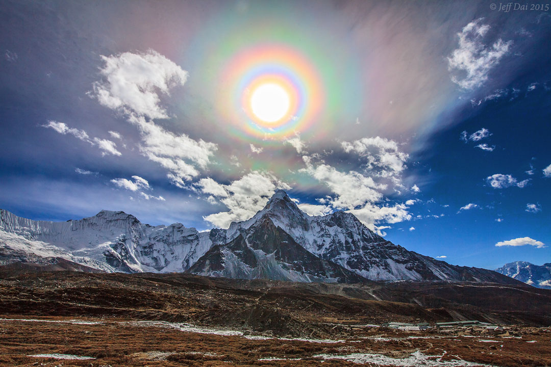 Über einem teils schneebedeckten Gebirge leuchtet die Sonne, sie ist von vielen bunten Ringen umgeben. Dieser Hof entsteht durch einen quantenmechanischen Effekt.