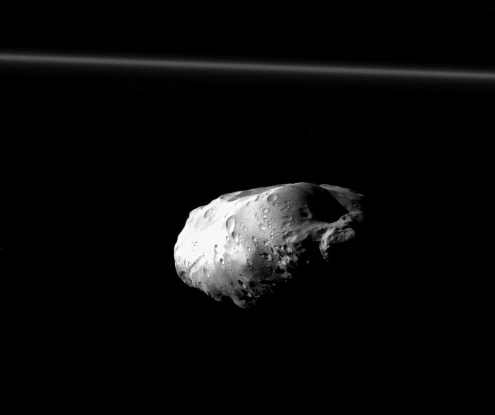 Der von Kratern übersäte Mond Prometheus schwebt mitten im Bild, oben verläuft der dünne F-Ring im Hintergrund.