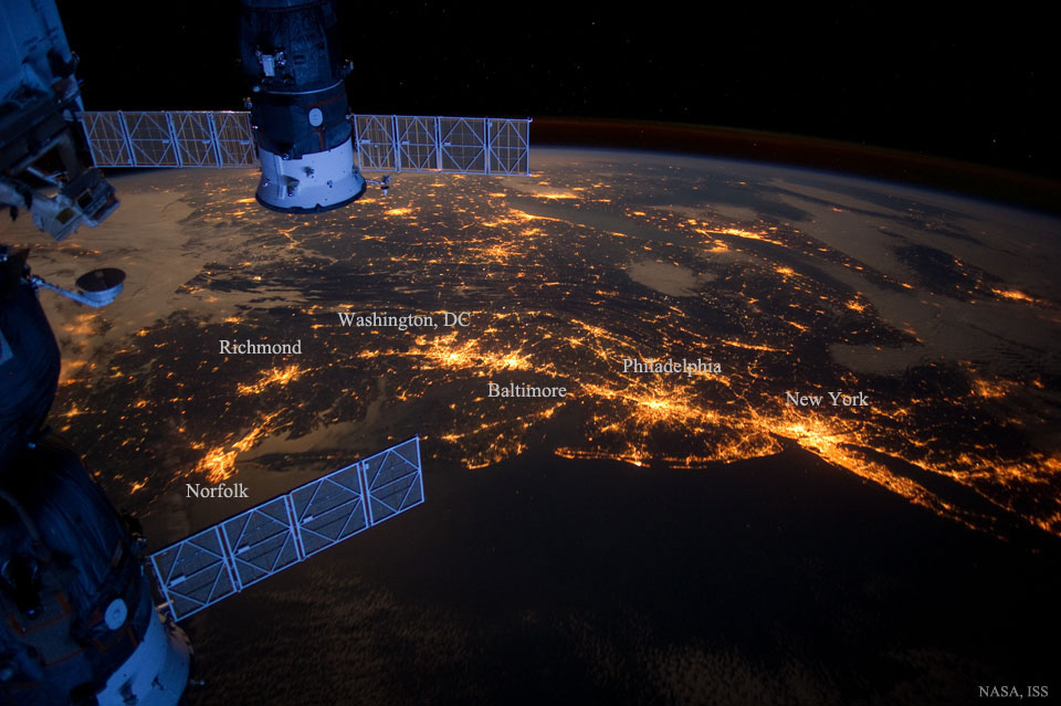 Der Blick fällt von der Raumstation auf die Erde. Unten ist das dunkle Meer, an der Küste sind Städte an der Ostküste der USA. Links sind Teile der Raumstation und Raumkapseln zu sehen.