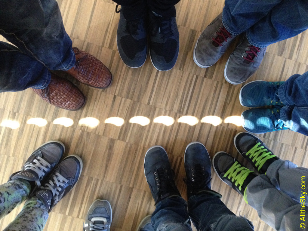 Auf einem Parkettboden stehen mehrere Leute mit Schuhen. Dazwischen verläuft eine Perlenkette mit Sonnenbildern, die angeknabbert sind. Es sind Lochkamerabilder der teilweise verfinsterten Sonne.
