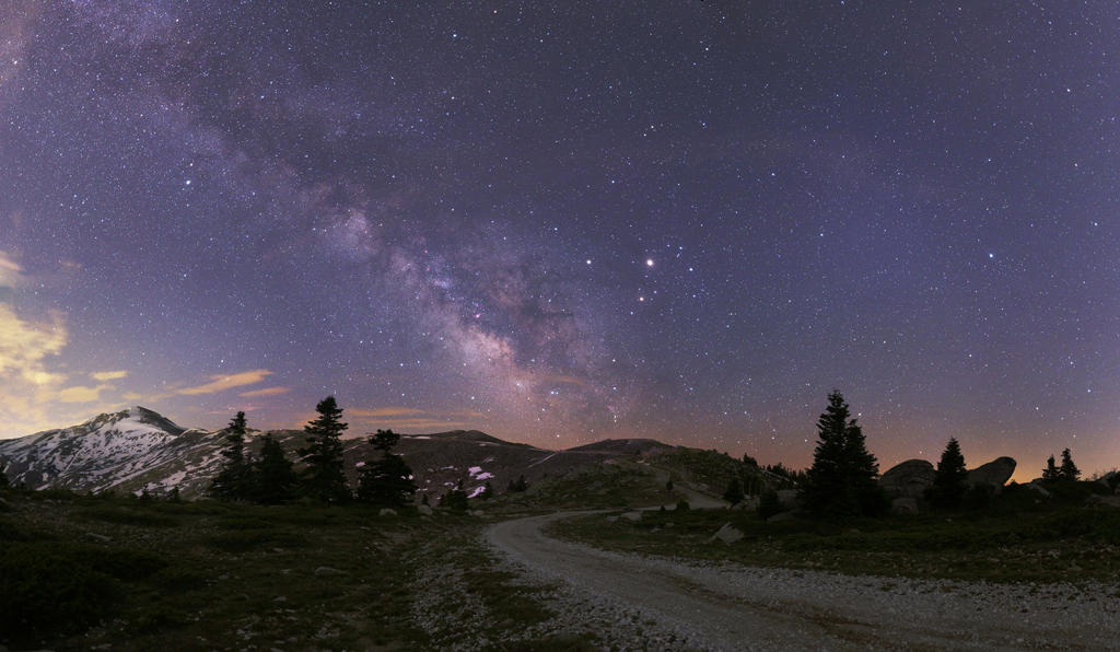 Eine Straße führt zu Planeten und der Milchstraße am Himmel. Links am Horizont sind verschneite Berggipfel.