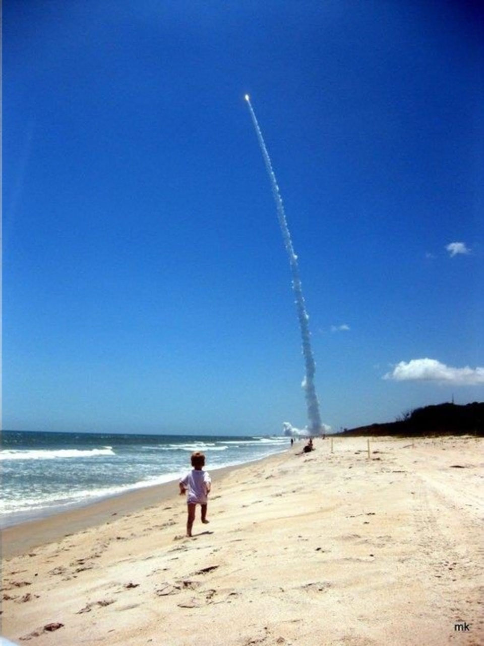 Ein Kind läuft einen Sandstrand entlang in Richtung einer startenden Rakete. Die Rakete bringt die Mission Juno ins All.