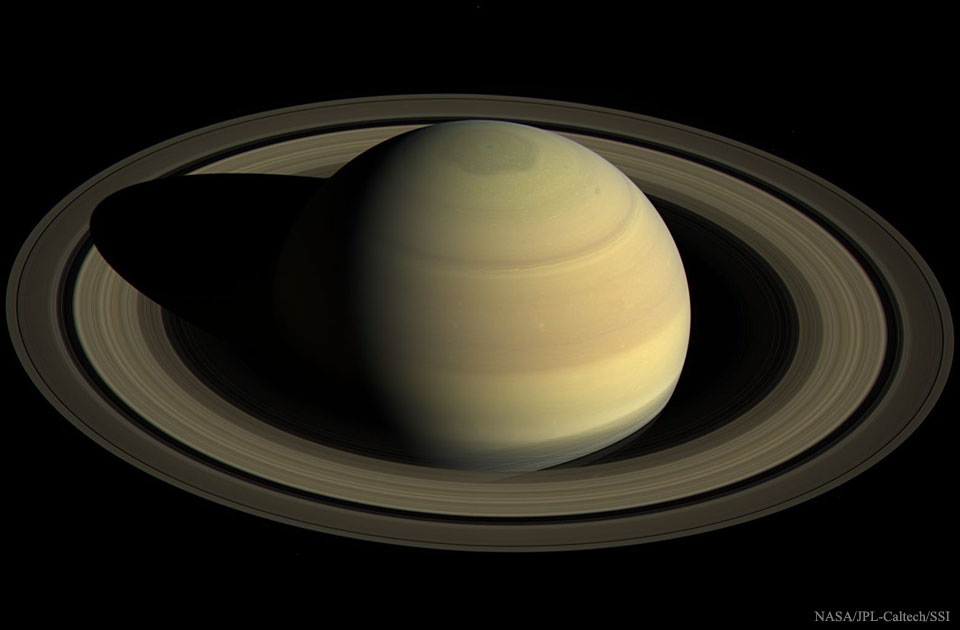 Saturn ist von schräg oben zu sehen, die Ringe füllen von links nach rechts die ganze Bildbreite. Am Pol des goldbraun beleuchteten Planeten ist eine sechseckige Wolkenstruktur.