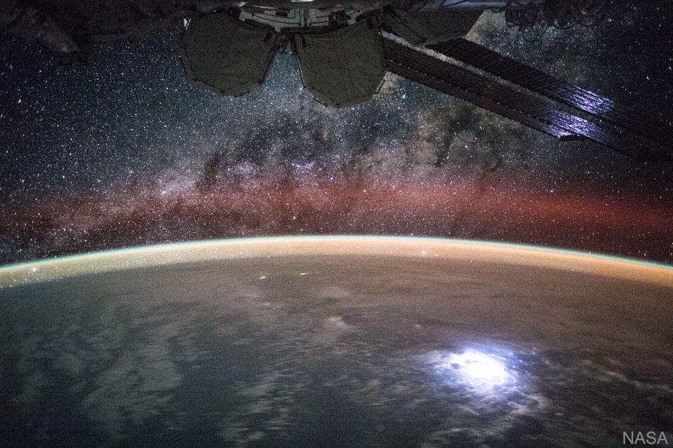 Die ISS zieht über die Erde. Unten blitzt ein Gewitter unter Wolken. Am Horizont ist braunes und grünes Nachthimmellicht. Am Himmel schimmert die Milchstraße.
