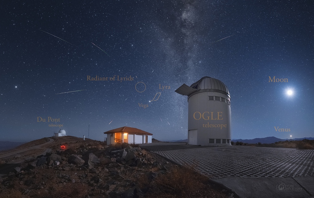 Hinter den Kuppeln und Gebäuden des Las Campanas Observatoriums steigt in der Bildmitte die Milchstraße auf. Rechts leuchten Mond und Venus. In der Mitte ist das Sternbild Leier und der Radiant der Lyriden, die durchs Bild flitzen.