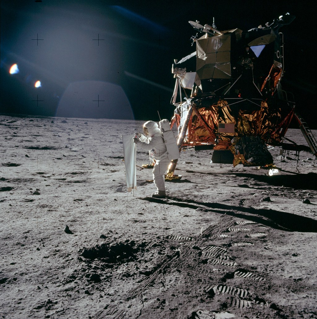 Rechts hinten steht das Mondlandemodul "Adler". Davor in der Mitte rollt Buzz Aldrin eine Folie aus und richtet sie zur Sonne. Sein langer Schatten fällt nach rechts über den dunkelgrauen Mondboden.