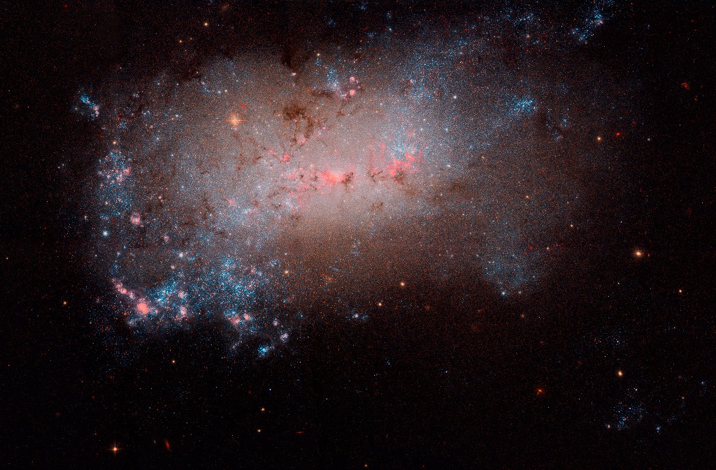 Die irreguläre Galaxie im Bild ist von vielen rötlich und bläulich leuchtenden Sternhaufen überzogen.