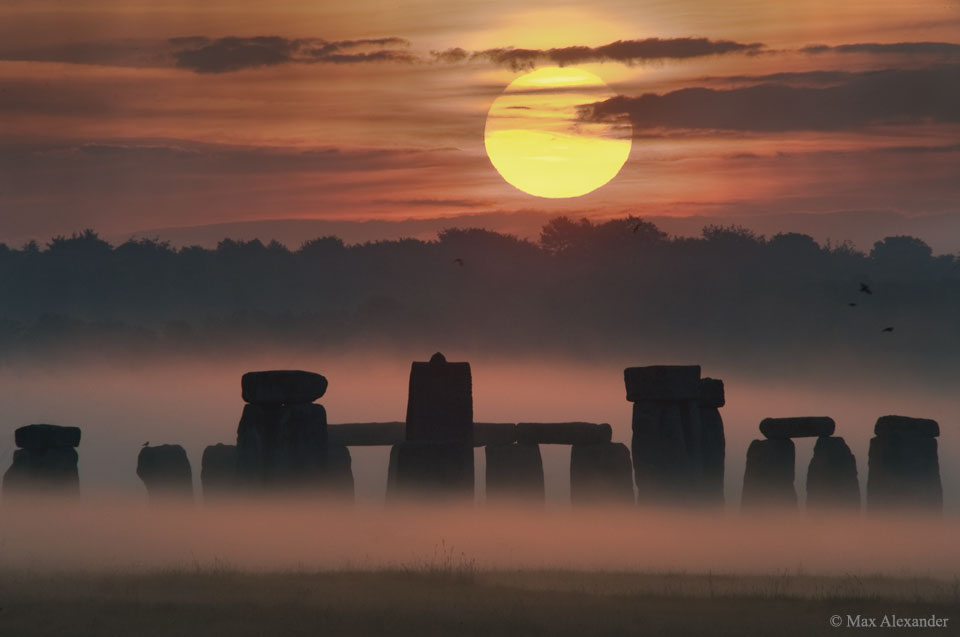 Sonnenaufgang zur Sommersonnenwende 2008 bei Stonehenge in Großbritannien. Die Sonne ist teilweise von Wolken verdeckt, die Wolken bilden dunkle waagrechte Bänder am orangefarbenen Himmel.