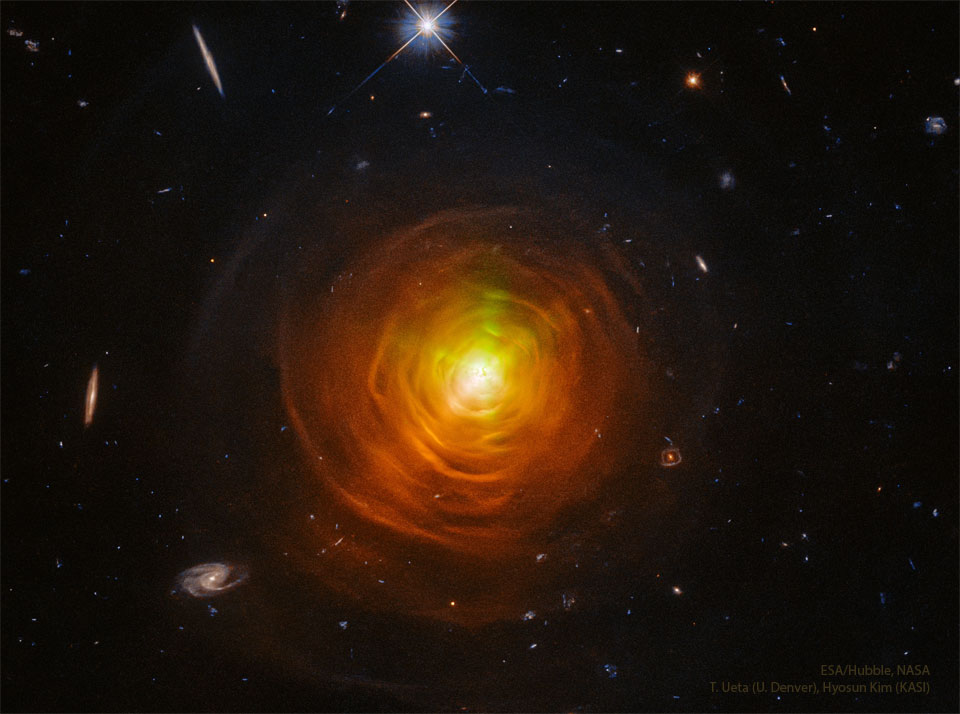 In der Mitte leuchtet ein Stern, der von vielen Hüllen um geben ist, die als Bögen abgebildet sind. Links im Bild sind drei Galaxien erkennbar, zwei davon sehen wir fast von der Kante. Im ganzen Bild sind weitere kleinere Galaxien verteilt.