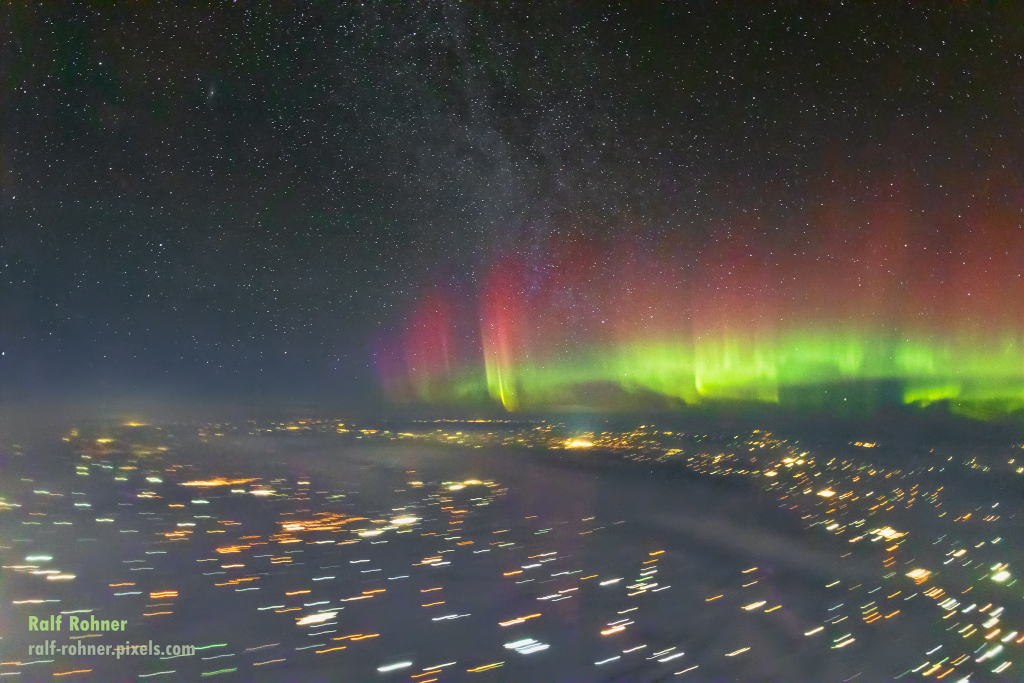 Rechts über dem Horizont leuchten Polarlichter, unten grün und darüber dunkelrot. Über dem Horizont sind zart gesprenkelte Sterne, unten auf der Erde sind zahlreiche Lichtspuren.