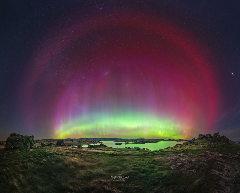 Über einer Landschaft mit einem Gewässer leuchtet ein Polarlicht. Über dem Horizont ist ein intensives, helles grünes Licht, darüber ein viel dunkleres purpurnes Leuchten.