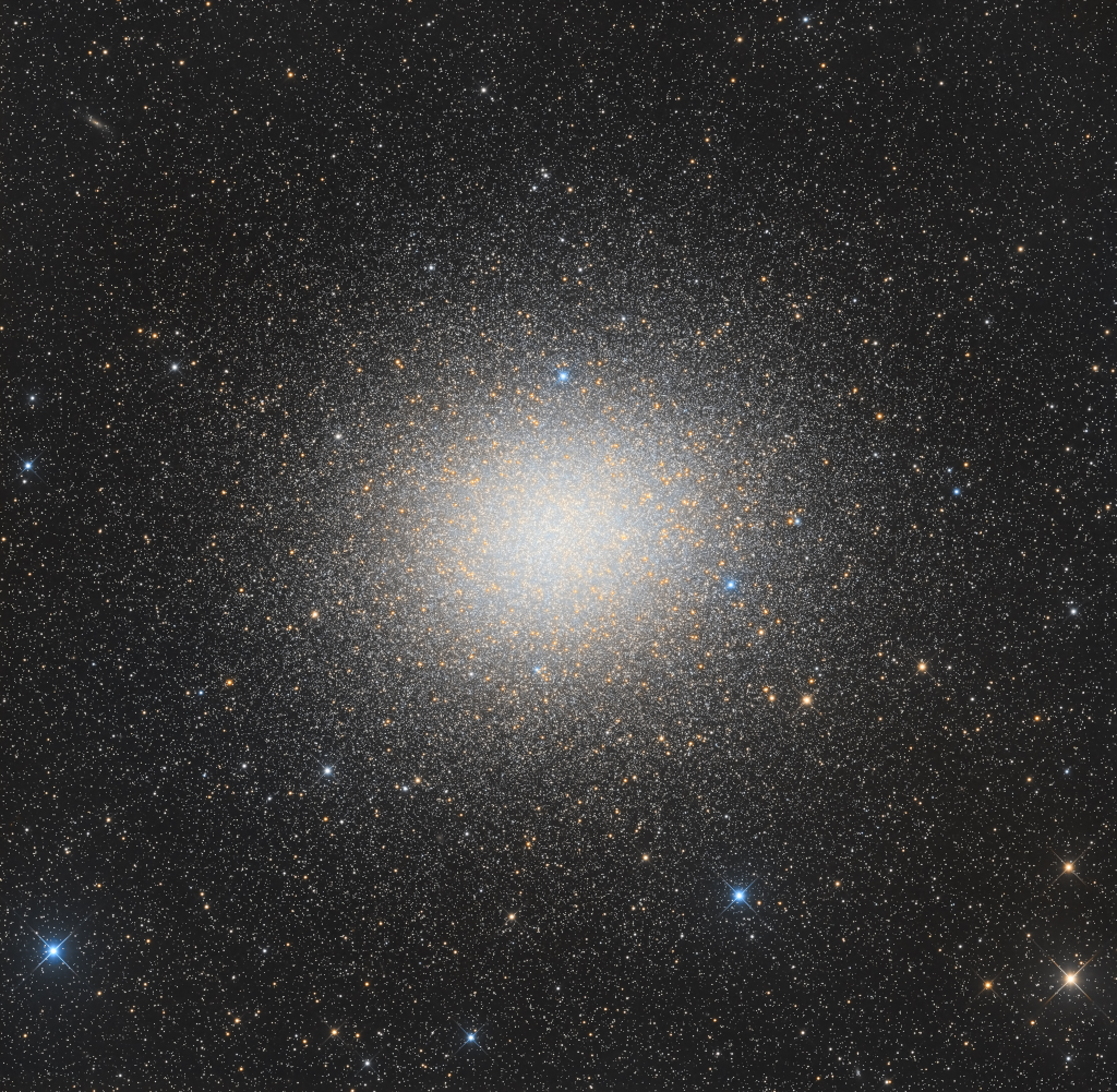 Der Kugelsternhaufen im Bild ist riesig und sehr dicht voller Sterne. Im Sternhaufen sind zahlreiche gelblich leuchtende Sterne verteilt.