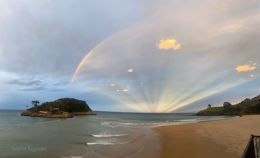 Über einem Strand in Spanien breitet sich am Horizont ein Fächer aus Gegendämmerungsstrahlen aus. Die Strahlen wird von einem Regenbogen am wolkigen Himmel begrenzt.