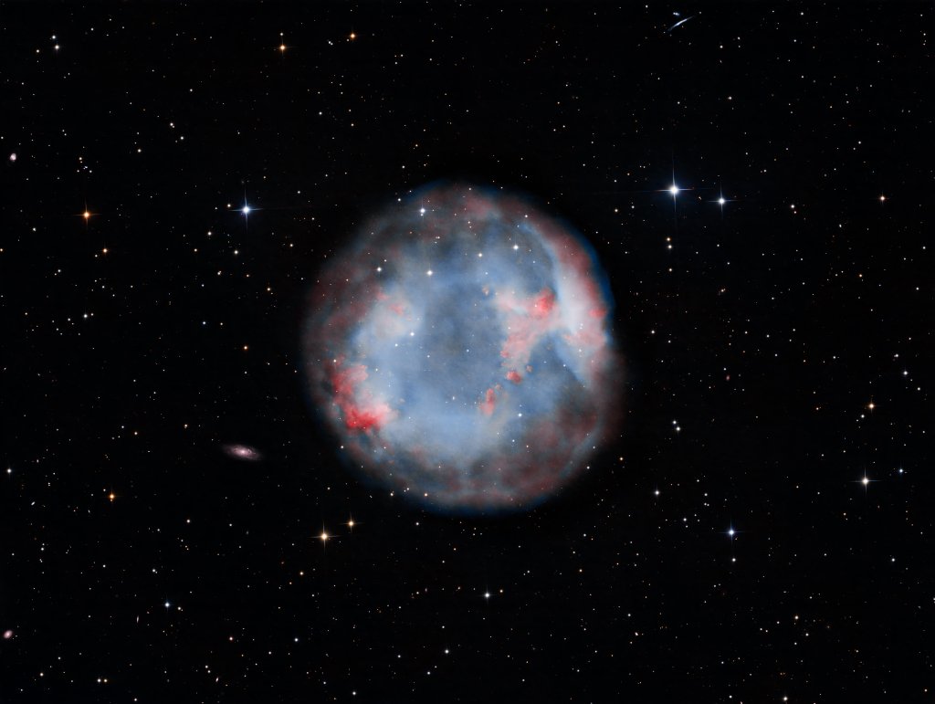 Ein kugelförmiger, blau-grauer Nebel leuchtet mitten im Bild. Er ist von roten Sprenkel überzogen und von wenigen Sternen umgeben. Ein bisschen erinnert er an die Iris in einem Auge.