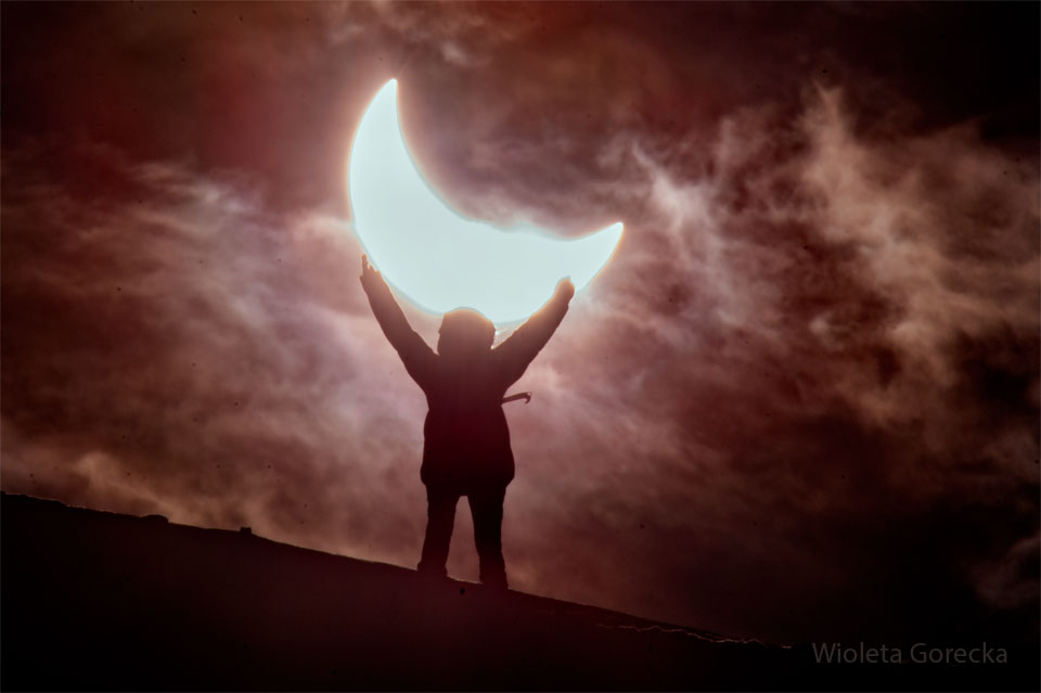 Eine Person hält scheinbar die Sonne, die nur als Sichel zu sehen ist, weil sie oben vom Mond verdeckt wird. Die Person ist als Silhouette zu sehen, der Himmel ist von Wolken verdeckt.