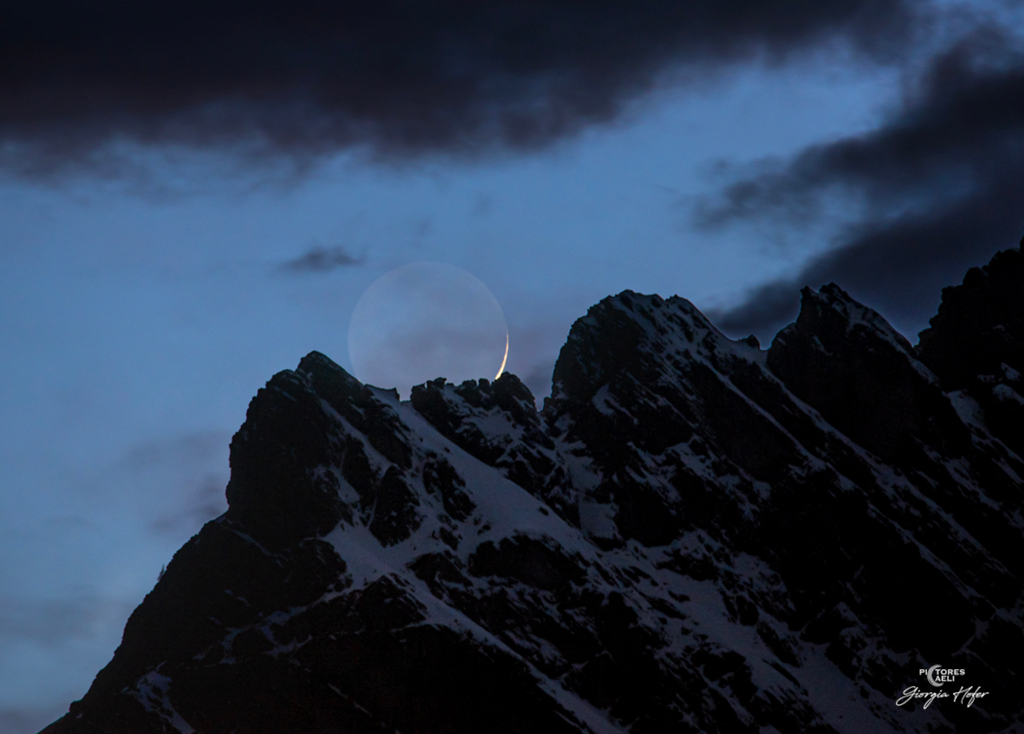 Über den italienischen Dolomiten geht ein schmaler Sichelmond auf. Die dunkle Nachtseite ist von Licht beleuchtet, das die Erde reflektiert.