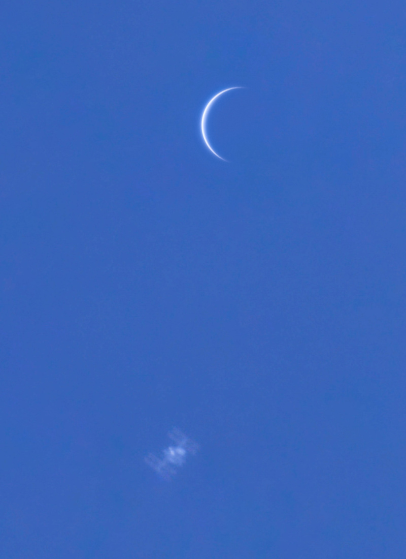 Am blauen Tageshimmel leuchtet oben die schmale, sichelförmige Venus, unten ist die verschwommene Silhouette der Internationalen Raumstation mit Solarpaneelen und Modulen zu sehen.