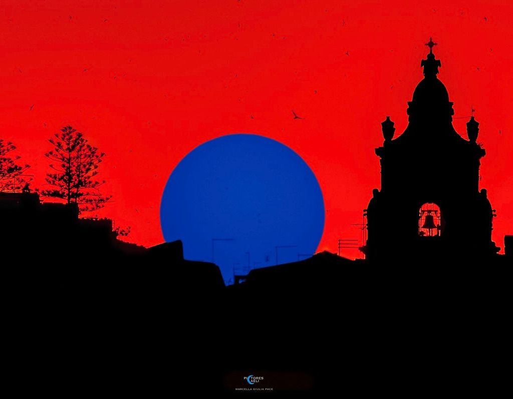 Hinter der Silhouette, die einen Kirchturm in Ragusa auf Sizilien zeigt, geht eine blaue Sonne auf einem roten Himmel unter. Wenn man das Bild länger betrachtet und dann die Augen schließt, sieht man ein Nachbild in natürlichen Farben.