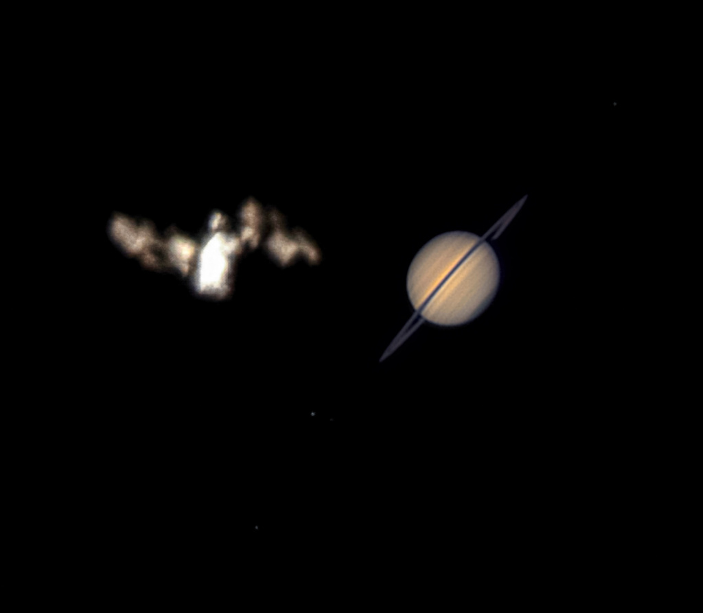 Neben dem Planeten Saturn, der rechts mit schmalen Ringen zu sehen ist, sieht man links die Internationale Raumstation ISS als verschwommene Gestalt.