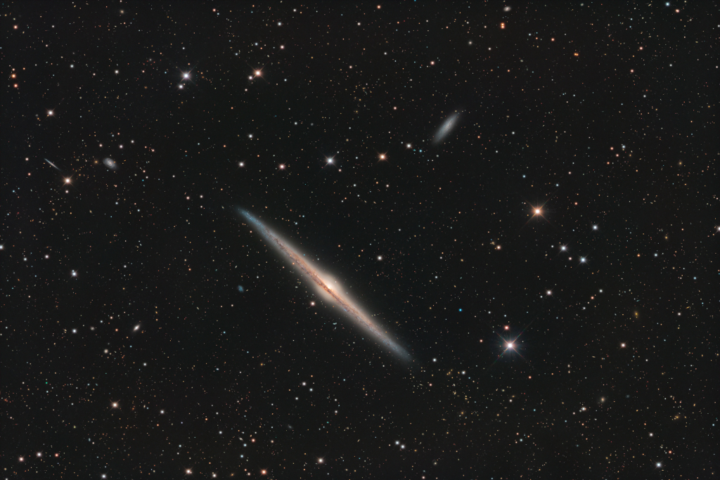 Die Galaxie NGC 4565 liegt diagonal mitten im Bild, sie ist von Sternen und Galaxien umgeben. Wir sehen auf ihre Kante, die von einem Staubrand gesäumt ist.