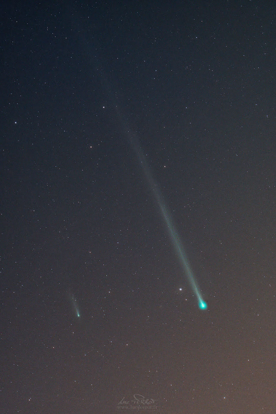 Zwei Kometen ziehen nebeneinander über den Himmel. Von ihren grünlichen Köpfen zeigt ein Ionenschweif nach links oben. Der Schweif des rechten Kometen ist viel länger als der des linken. Er ist auch viel näher.