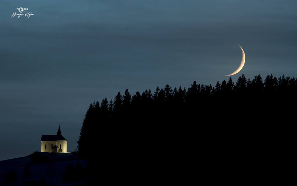 Über einem Nadelwald geht der Sichelmond in der Abenddämmerung unter. Links neben dem Wald steht eine kleine Kirche in der Nähe von Danta di Cadore in Norditalien.