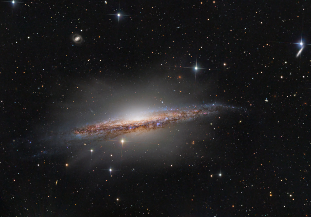 Die Galaxie NGC 1055 ist fast von der Kante zu sehen. Sie enthält viel dunklen Staub und ist von einem zarten Halo aus Sternen umgeben. Dahinter sind einige weitere Galaxien verteilt. Der Vordergrund ist lose mit unterschiedlich hellen Sternen dekoriert.
