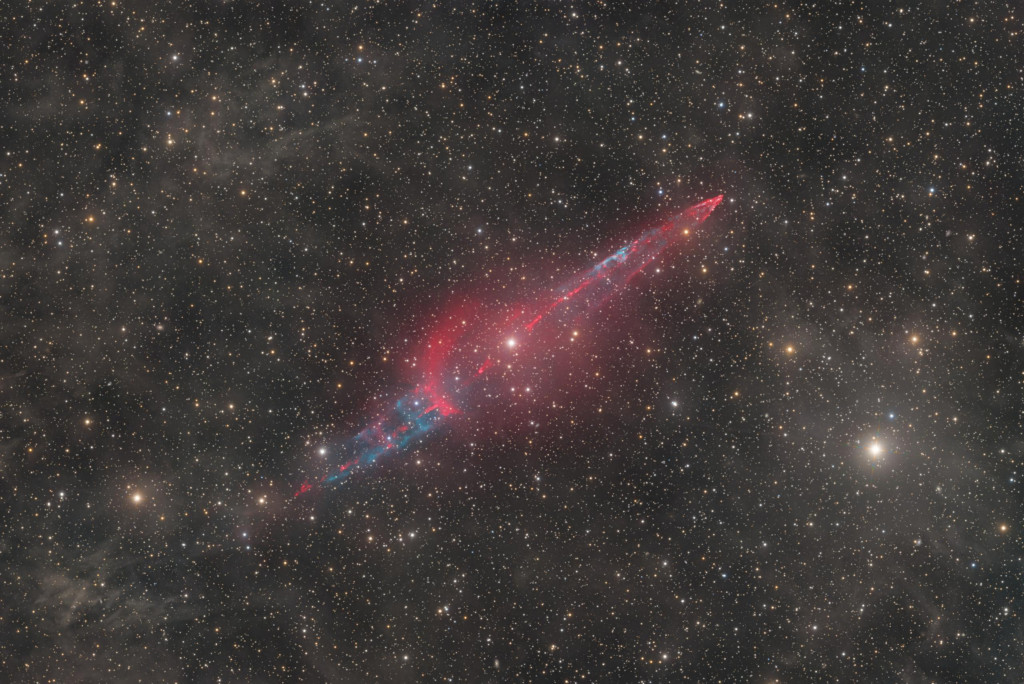 Aus dem Doppelstern KX Andromedae mitten im Bild schießen Strahlströme, die erst kürzlich entdeckt wurden. Sie sind leuchtend rot abgebildet. Das Bild ist von zarten diffusen Nebeln und dicht verteilten Sternen gefüllt.