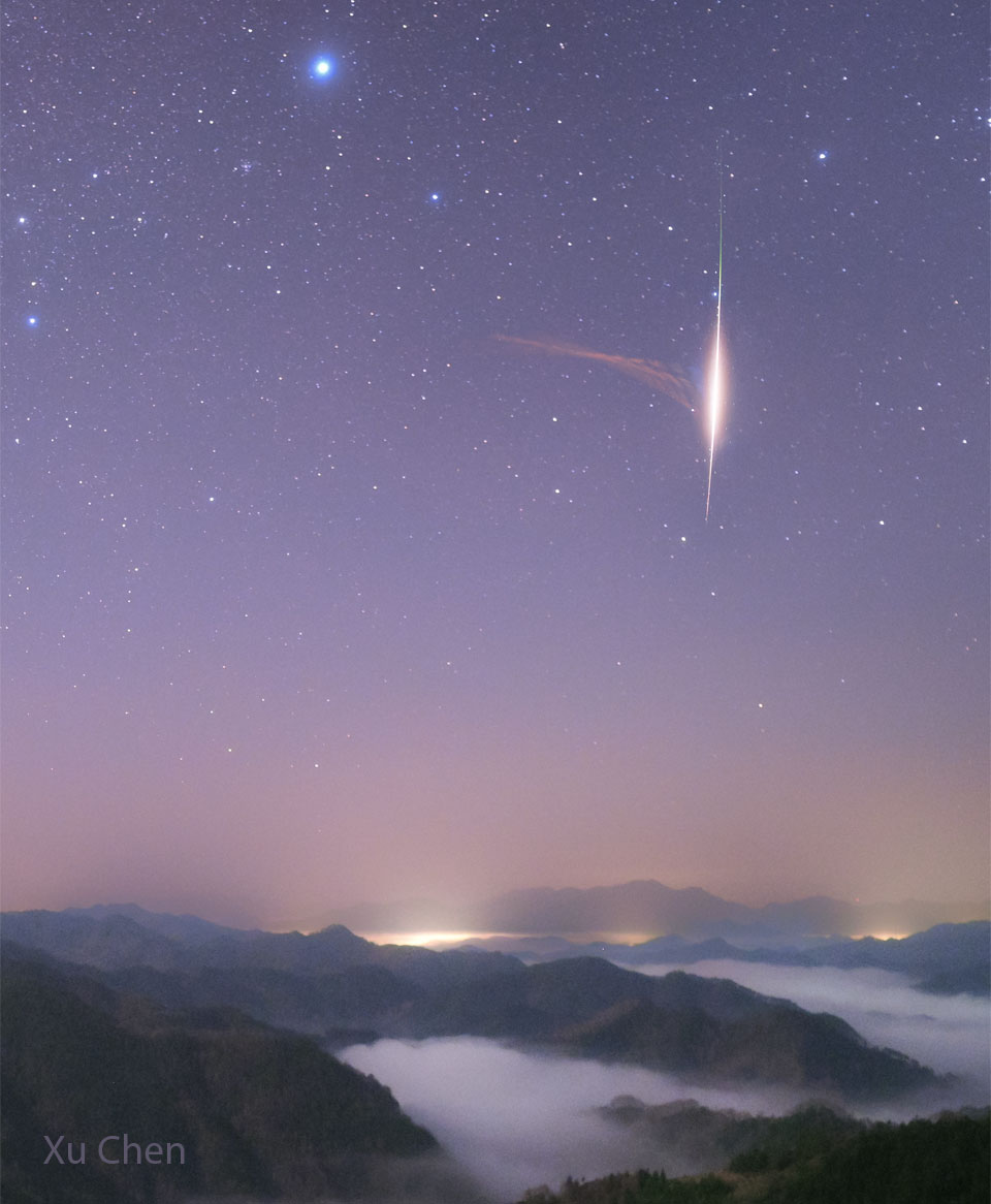 Über den nebelverhangenen Gelben Bergen (Huang Shan) im Osten von China flammt ein heller Meteor der Geminiden am sternklaren Himmel auf. Nach links zieht eine rötliche blasse Staubspur. Oben strahlt der helle Stern Sirius.