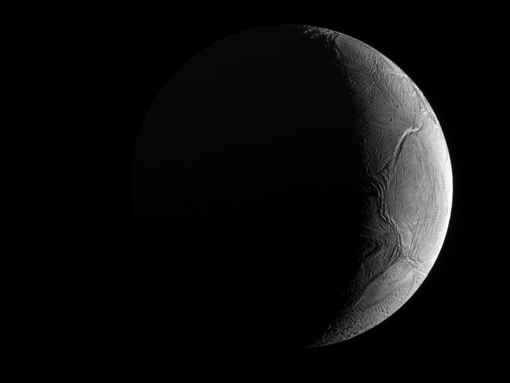 Rechts ragt die Sichel des Saturnmondes Enceladus, die von der Sonne beleuchtet wird, ins Bild. Die linke Seite ist dunkel. Die Oberfläche des Eismondes ist von tiefen Rillen überzogen. Unten sind auch Krater erkennbar.