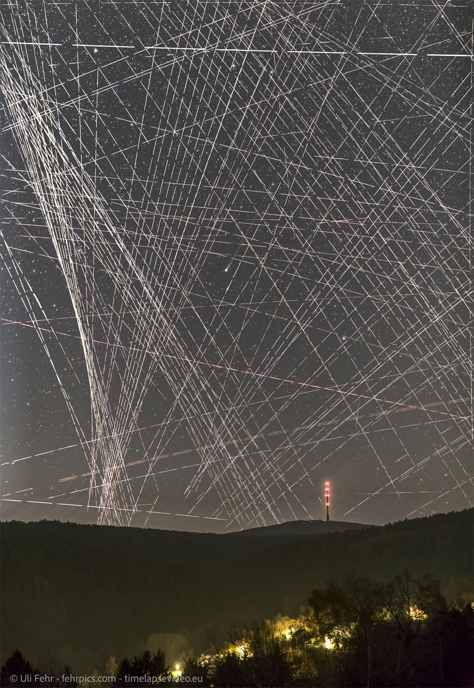 Über einem Horizont, auf dem ein rot beleuchteter Mast steht, ziehen Satelliten und Flugzeuge kreuz und quer helle Striche. Der Komet schimmert mittendrin, doch hinter den Spuren ist er kaum erkennbar. Rechts über der grünlichen Koma ragt sein schmaler Schweif leicht nach rechts.