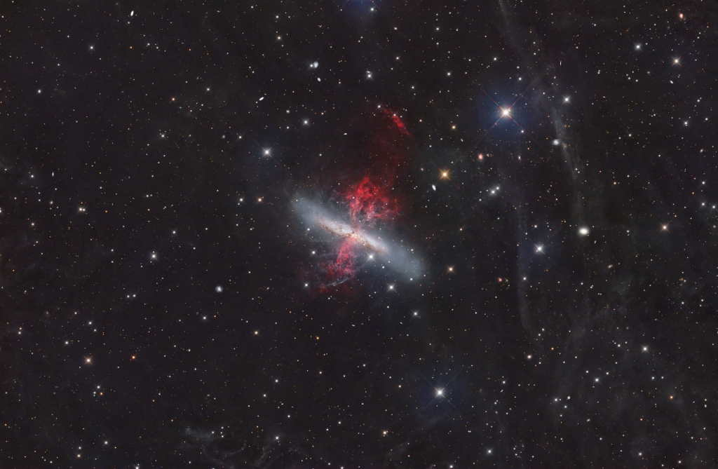 Die längliche Galaxie Messier 82 im Sternbild Große Bärin ist von einem Superwind umgeben. Aus der Mitte strömen rote Gasfäden, außen herum erkennt man zarte Schleier. Im Hintergrund sind lose Sterne verteilt.