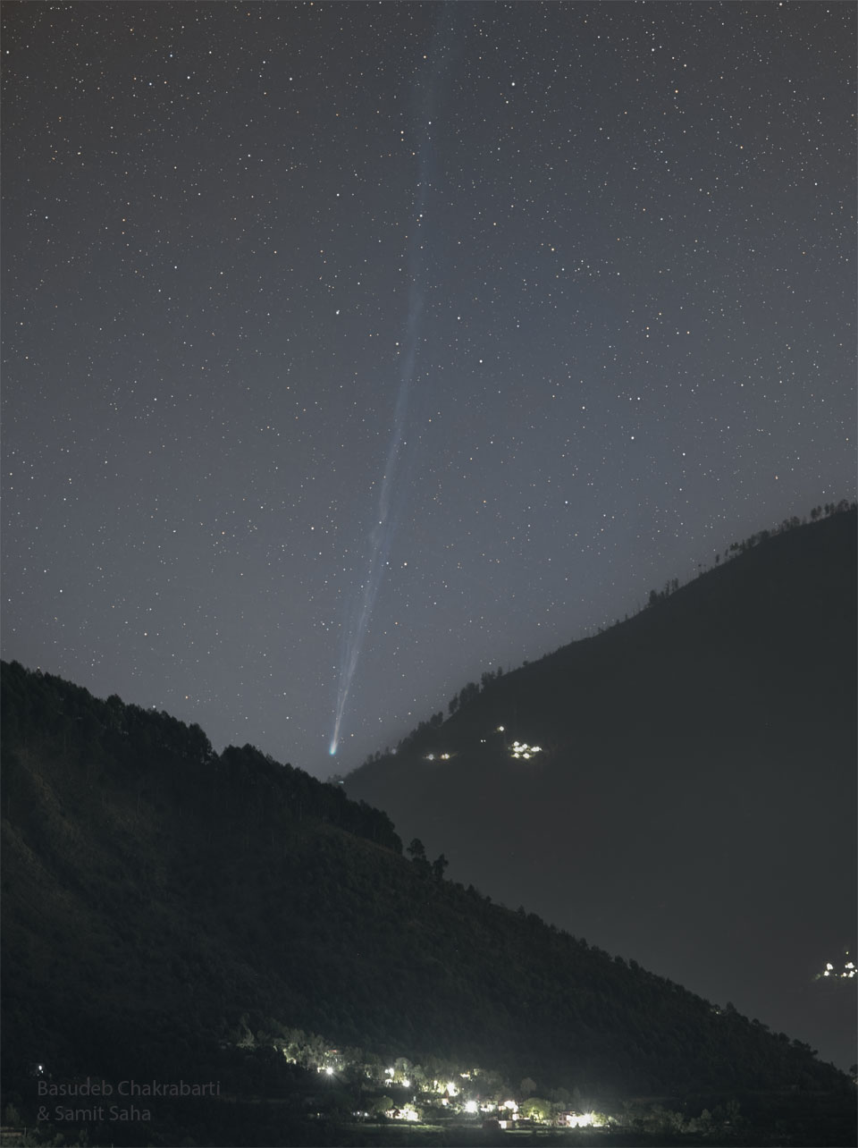Über einer Kerbe zwischen zwei Hängen, die nach links und rechts aufragen, steigt der Komet PanSTARRS mit seinem langen schmalen Schweif auf. Unten sind die Lichter einer Siedlung.