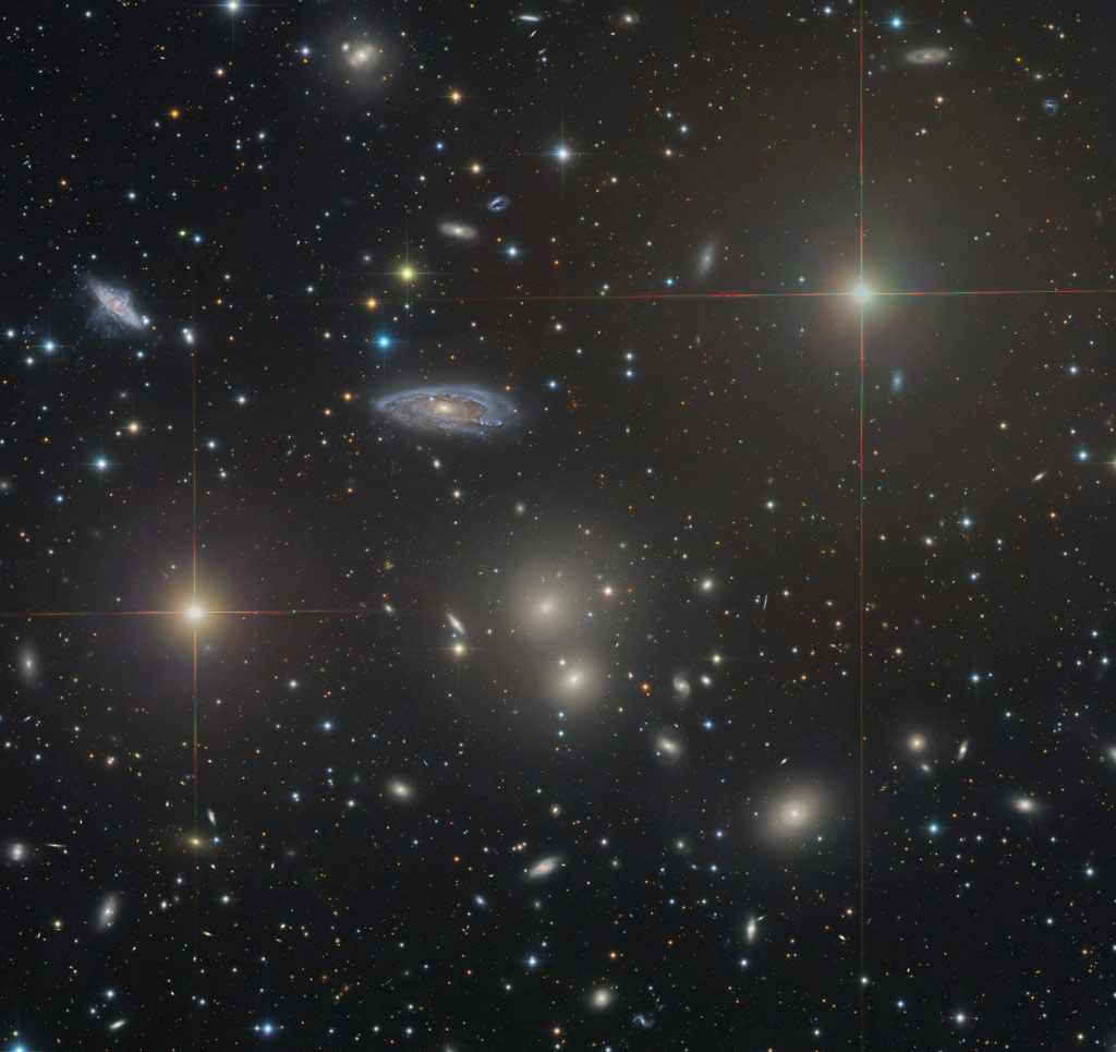 Links unten und rechts oben strahlen zwei helle Sterne mit Zacken. Dahinter sind viele Galaxien verteilt, etliche davon sind sehr klein.
