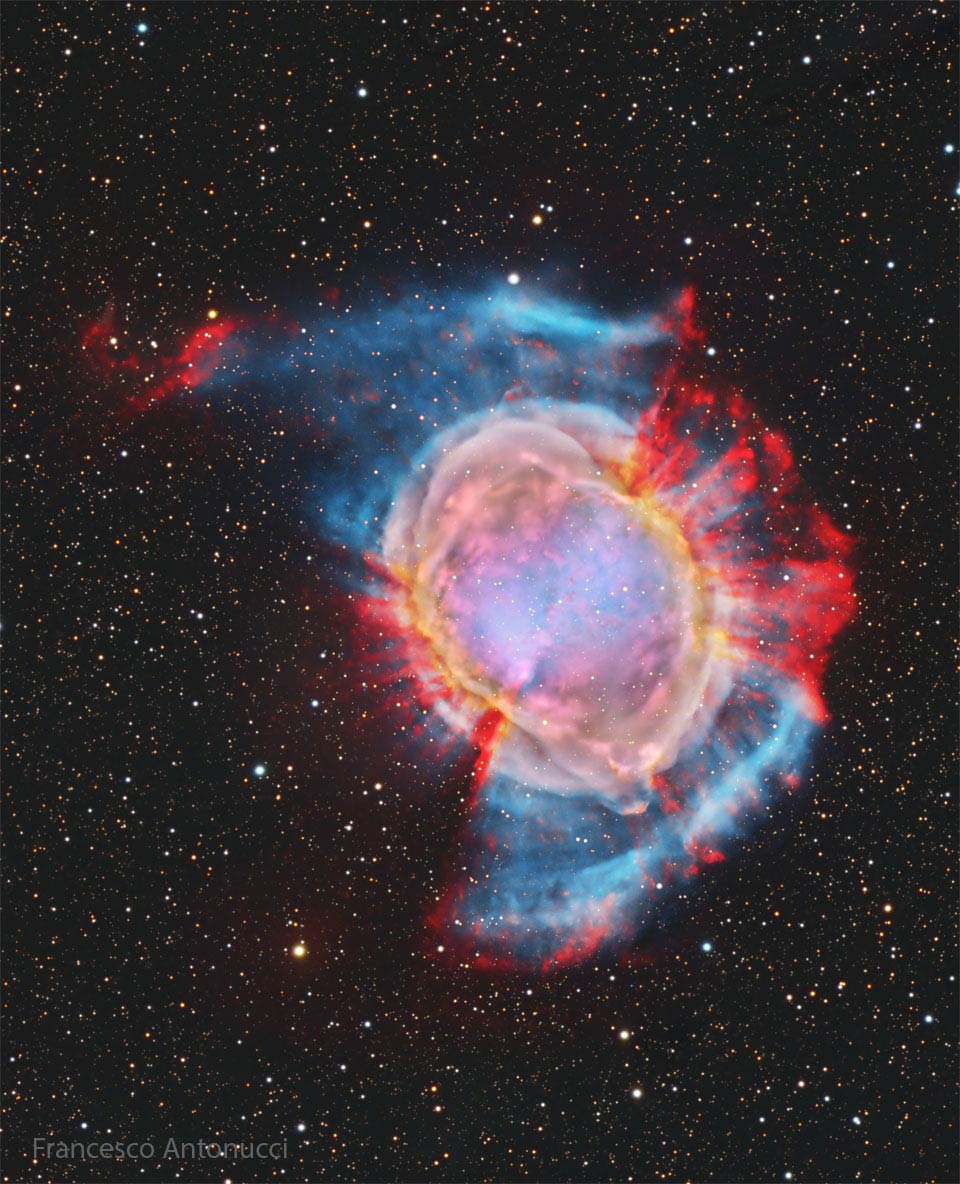 Ein großer, farbenprächtiger Nebel ist von einem Sternfeld umgeben. Außen ragen in einigen Richtungen rote Strahlen aus dem Nebel. An anderen Rändern zieht ein blauer Schleier. Das Zentrum des Nebels ist bunt.