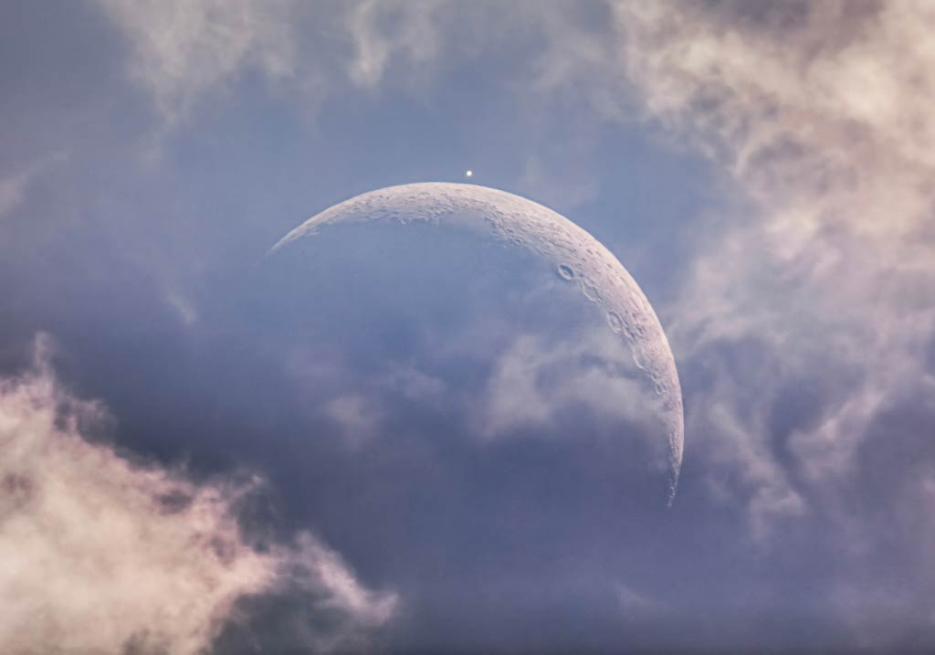 Hinter dramatischen Wolken leuchtet der von Kratern übersäte Sichelmond am blauen Himmel. Oben gleißt ein heller Lichtpunkt. Es ist die Venus, die bald vom Mond verdeckt wird.