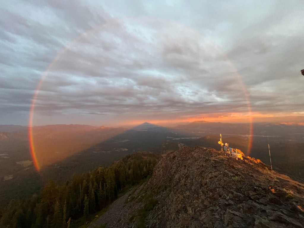 Ein Berg wirft einen dreieckigen Schatten, dessen Spitze am Horizont liegt. Rund um die Spitze des Schattengegels verläuft ein kreisförmiger Regenbogen. Der Himmel ist bewölkt, der rötliche Sonnenschein verleiht dem Bild einen etwas schaurigen Eindruck.